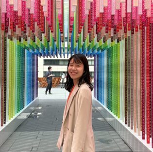 カラフルな数字の短冊が並ぶ通路で微笑む、ベージュのジャケットを着た女性。